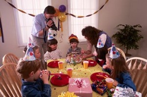 A father videotapes his son's birthday party as the mother brings the cake to the table.